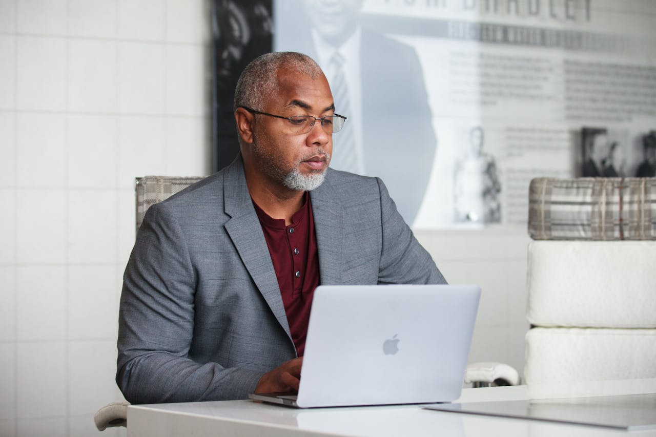 our-services-3 Senior adult man with gray beard working remotely on laptop indoors wearing a blazer.