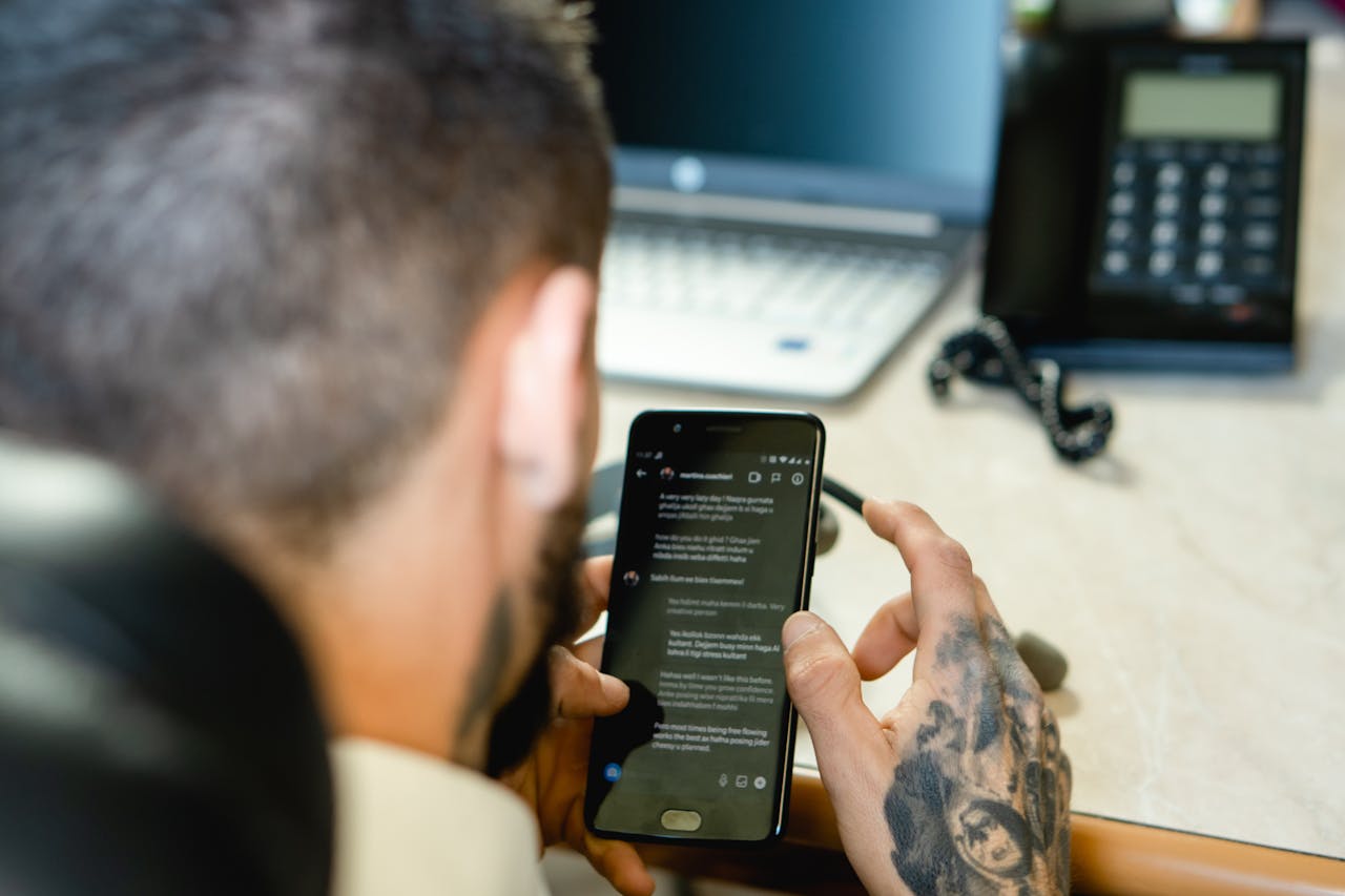 why-choose-us A tattooed man uses a smartphone at a desk with a laptop and a phone, symbolizing modern communication.